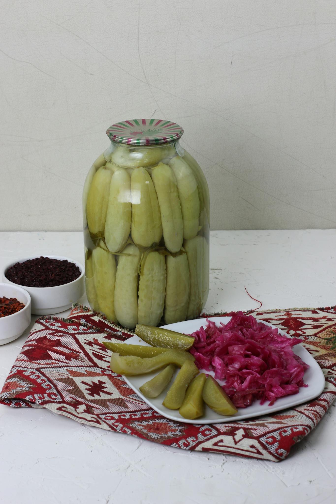 A jar of freshly pickled cucumbers with red cabbage plated on a patterned cloth, indoors setup.